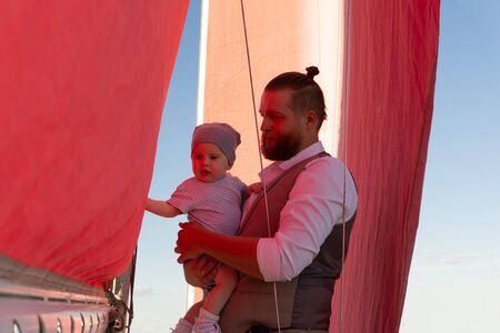 bearded man holding a baby in his arms against the background of blue sky and a ship mast with red...