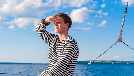 young male yachtsman is looking away from under his arm against the background of water space with a distant shoreの写真素材