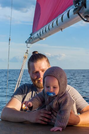 bearded man holding a baby in his arms against the background of blue sky and a ship mast with red sailsの写真素材