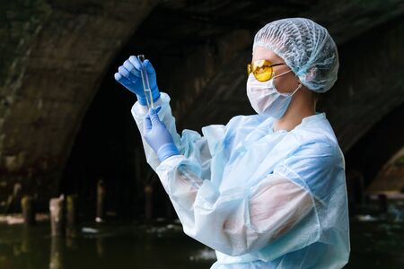 female ecologist or biochemist examines a test tube with water from a city river in an underground...