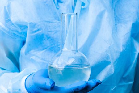 researcher's hand in a glove holds a glass laboratory flask with liquid, background in blue tones