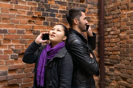 young man and woman stand back to back near a brick wall talking on their cell phones