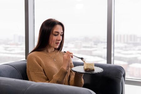 girl eating a cheesecake while sitting in a chair in front of a large window on a winter dayの写真素材