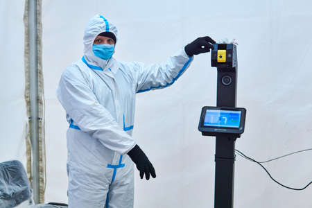 Perm, Russia - April 25, 2020: medical officer in protective suit invites to take a non-contact temperature measurement in the tent of the sanitary and epidemiological control point installed at the railway stationのeditorial素材