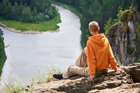 teenager sits on the edge of a cliff looking at the river against the backdrop of a natural landscapeの写真素材