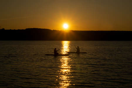 two surfers sitting and row on standup paddleboards against the backdrop of the setting sunの写真素材