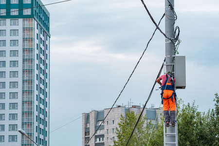 Perm, Russia - August 25, 2021: electrician or telecommunications lineman works on laying a cable at the top of a telephone pole against the background of city buildingsのeditorial素材
