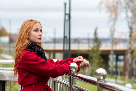 beautiful red-haired girl looks into the distance while standing over the autumn city embankmentの写真素材