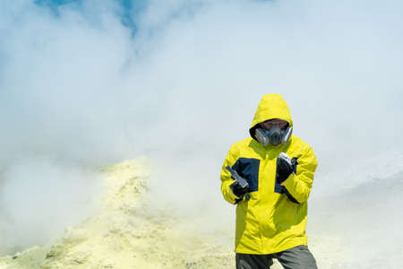 male volcano scientist on the slope of a volcano among the vapors of a fumarole examines a sample of a mineralの写真素材