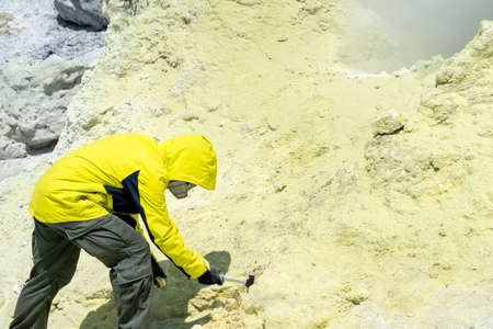 male volcanologist on the slope of a volcano next to a smoking fumarole takes a sample of a mineral from the edge of a sulfurous craterの写真素材
