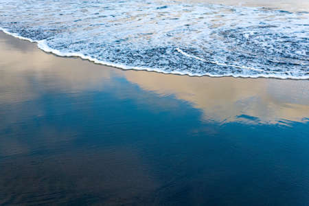 ocean beach with black volcanic sand, the sky is reflected in the rolled back wave of the surf