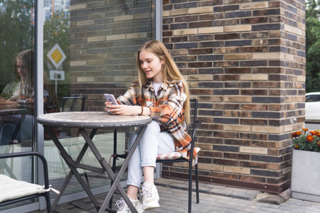 young woman reading funny message on phone while sitting at an outdoor cafeteria tableの写真素材