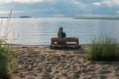 woman traveler with de braids hairstyle sitting on a bench made of old pallets on a sandy shore of a vast lakeの写真素材