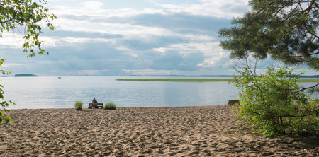 sandy beach on the shore of a vast lake, a female tourist is sitting on a bench made of old palletsの写真素材