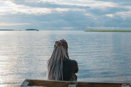 woman traveler with de braids hairstyle sitting on a sandy shore of a vast lakeの写真素材