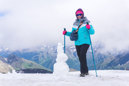 female mountaineer and snowman on the edge of a high mountain glacier against the backdrop of a green valley covered in cloudsの写真素材