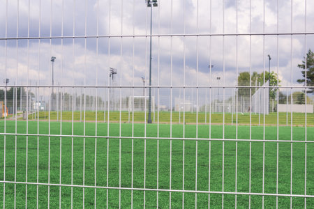Sports grounds and soccer fields behind a metal fence with green grass and blue skyの写真素材