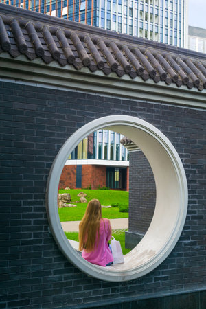 young woman sits in a round window in the fence of a Chinese garden
