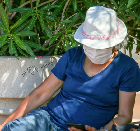 A woman wearing a hat and medical mask sitting on a bench. Cannes, August 2020.のeditorial素材