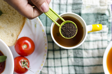 hand holding healthy black tea in table spoon with breakfast plate bread sauce and tomatoes backgroundの写真素材