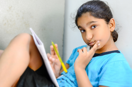 Indian Asian Girl child student writing & reading indoor doing homework sitting in room looking at cameraの写真素材