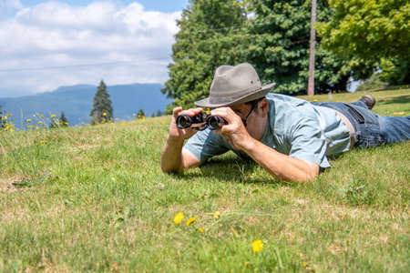 A side-view of a man lying down on a grass field and using binoculars.の写真素材