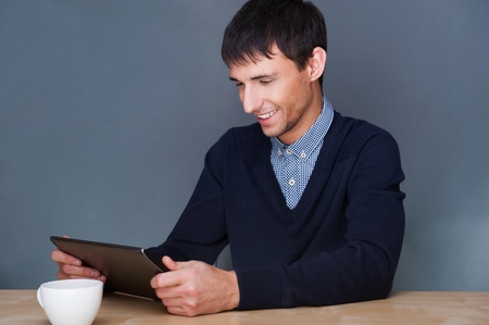 Closeup of a young smiling business man sitting at his office against gray wall and using his modern...