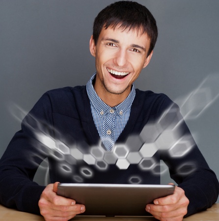 Closeup of a young smiling business man sitting at his office against gray wall and using his modern tablet pc to do his workの写真素材
