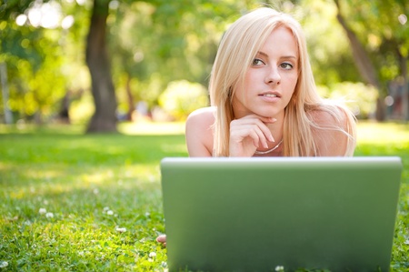 Portrait of young pretty woman resting on green grass at summer park using laptop to communiczte her friends. Outdoors. Daydreamingの写真素材