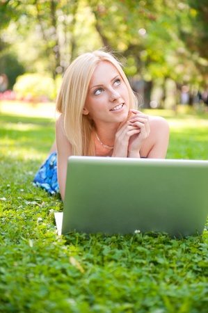 Portrait of young pretty woman resting on green grass at summer park using laptop to communiczte her friends. Outdoors. Daydreamingの写真素材