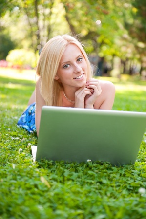 Portrait of young pretty woman resting on green grass at summer park using laptop to communiczte her friends. Outdoors. Daydreamingの写真素材