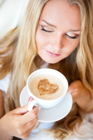 Smiling woman drinking a coffee lying on a bed at home or hotel. Heart shape illustrated on coffee foam. Vertical shot. Model is looking awayの写真素材