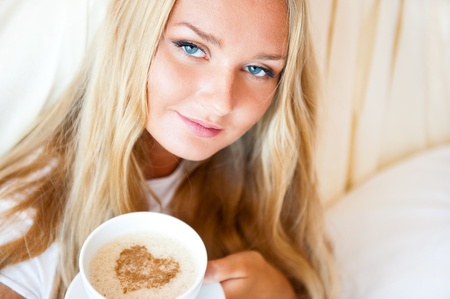 Smiling woman drinking a coffee lying on a bed at home or hotel. Heart shape illustrated on coffee foam. Horizontal shot. Model is looking to the camera. Lots of Copyspace.の写真素材