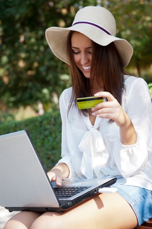 Young elegant woman wearing straw hat and white dress holding credit card with laptop sitting on bench at park and looking away. She is shopping or making payments carefree onlineの写真素材