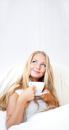 Smiling woman drinking a coffee lying on a bed at home or hotel. Daydreamingの写真素材