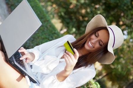 Young elegant woman wearing straw hat and white dress holding credit card with laptop sitting on bench at park and looking away. She is shopping or making payments carefree onlineの写真素材