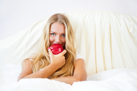 Closeup portrait of young pretty woman lying in her bed at morning and eating red appleの写真素材