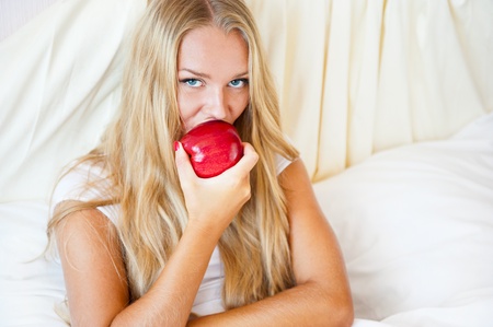 Closeup portrait of young pretty woman lying in her bed at morning and eating red appleの写真素材