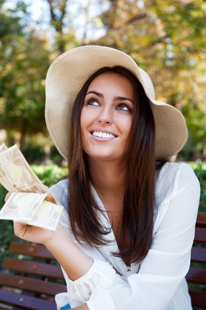 Portrait of young pretty woman sitting on bench at summer or autumn park and daydreaming while holding money and looking up to the sky. Wearing stylish clothes - straw hat and white blouseの写真素材