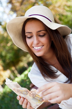 Portrait of young pretty woman sitting on bench at summer or autumn park and daydreaming while holding money and looking up to the sky. Wearing stylish clothes - straw hat and white blouseの写真素材