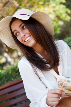 Portrait of young pretty woman sitting on bench at summer or autumn park and daydreaming while holding money and looking up to the sky. Wearing stylish clothes - straw hat and white blouseの写真素材