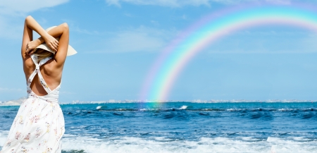 Young fashionable woman wearing hat and sophistical dress on the beach looking on rainbow. Photo from behindの写真素材