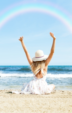 Portrait of young woman on the beach near the sea sitting with hands up wearing sophistical dress and hat looking at rainbow. Photo from behindの写真素材