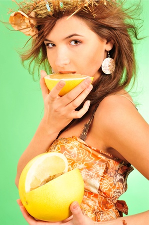 Closeup fashion portrait of pretty young woman wearing dress and headwear crown of hay and flower eating fresh pomelo fruit against beautiful green background. Freshness conceptの写真素材