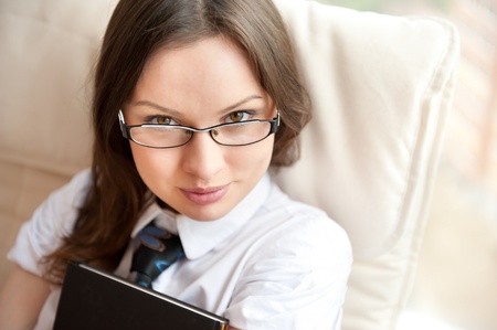Portrait of a happy young brunette woman holding book sitting on comfortable chair near window with natural light. Wearing eyeglassesの写真素材