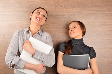 Portrait of two young handsome women standing relaxed against modern wooden wall in their office smiling and holding laptop and folder with papersの写真素材