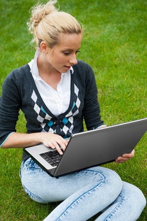 Young woman with laptop sitting on green grass and looking to the screen of laptopの写真素材