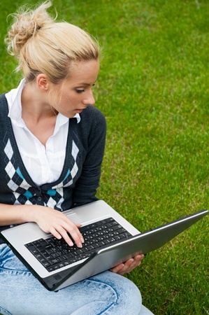 Young woman with laptop sitting on green grass and looking to the screen of laptopの写真素材
