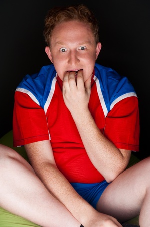 Closeup portrait of young man wearing sportswear fan of football team is watching tv and rooting for his favorite team. Sitting on beanbag alone at night. Light from television.の写真素材