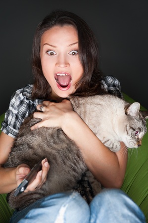 Young funny woman sitting in her beanbag and embracing her cat while watching tv. She is very scared while watching horror film in dark roomの写真素材